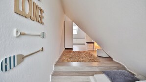 A narrow hallway with a wooden floor leading to a room with a white couch and a gray pillow. A wooden sign on the wall reads "Beach Lounge." - Domizil Gartenstraße 4 Appartement 7 Treibgut (Timmendorfer Strand)
