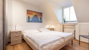 Un dormitorio con piso de madera, una cama con ropa blanca, un mesita de noche con lámpara, un cuadro de un muelle en la pared y una ventana con una planta en el alféizar.