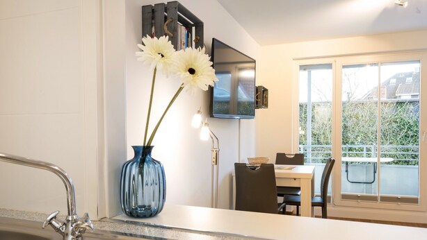 Una cocina moderna con un lavamanos de acero inoxidable, un florero azul con flores y una zona de comedor con mesa y sillas.