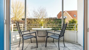 Un patio cubierto al aire libre con una mesa redonda y cuatro sillas, vistas a una zona residencial con árboles y casas, y una barandilla blanca.