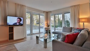 A modern living room with a flat-screen TV, a glass coffee table, a grey sofa with red cushions, and large windows offering a view of the outdoors. - Villa Stern Appartement 2 (Timmendorfer Strand)