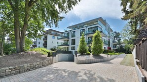 A modern multi-story building with a paved courtyard, surrounded by trees and a stone wall. - Villa Stern Appartement 2 (Timmendorfer Strand)