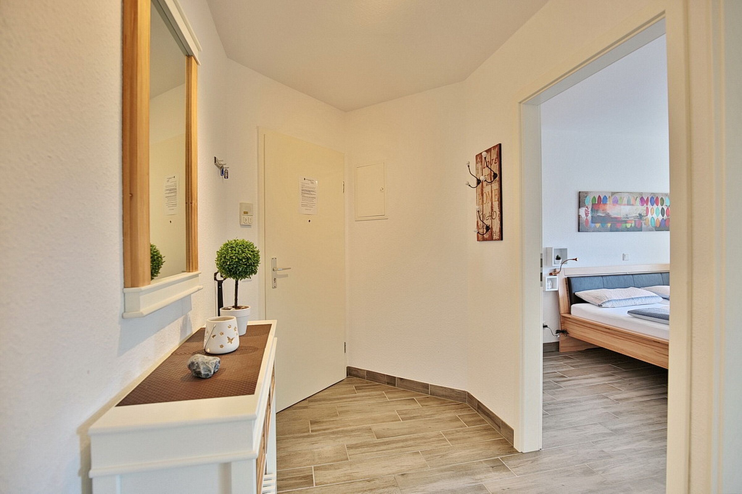 A hallway with a wooden bench, a small potted plant, and a mirror. There is a door on the left and a bed with a headboard in the background.