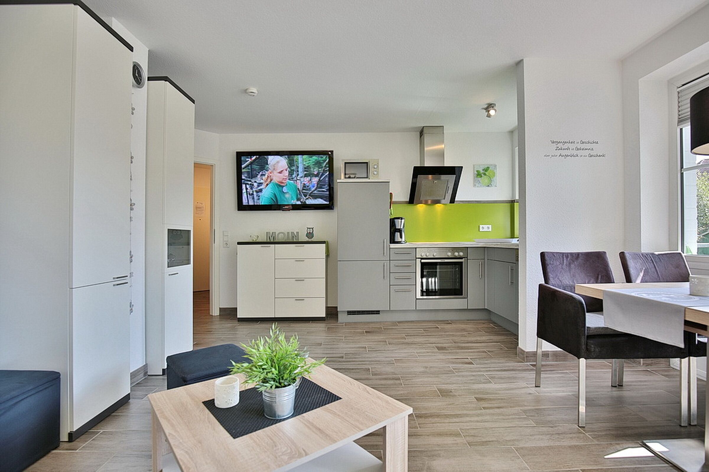 A modern living room with a television, a dining area with a table and chairs, and a kitchen with a green accent wall.