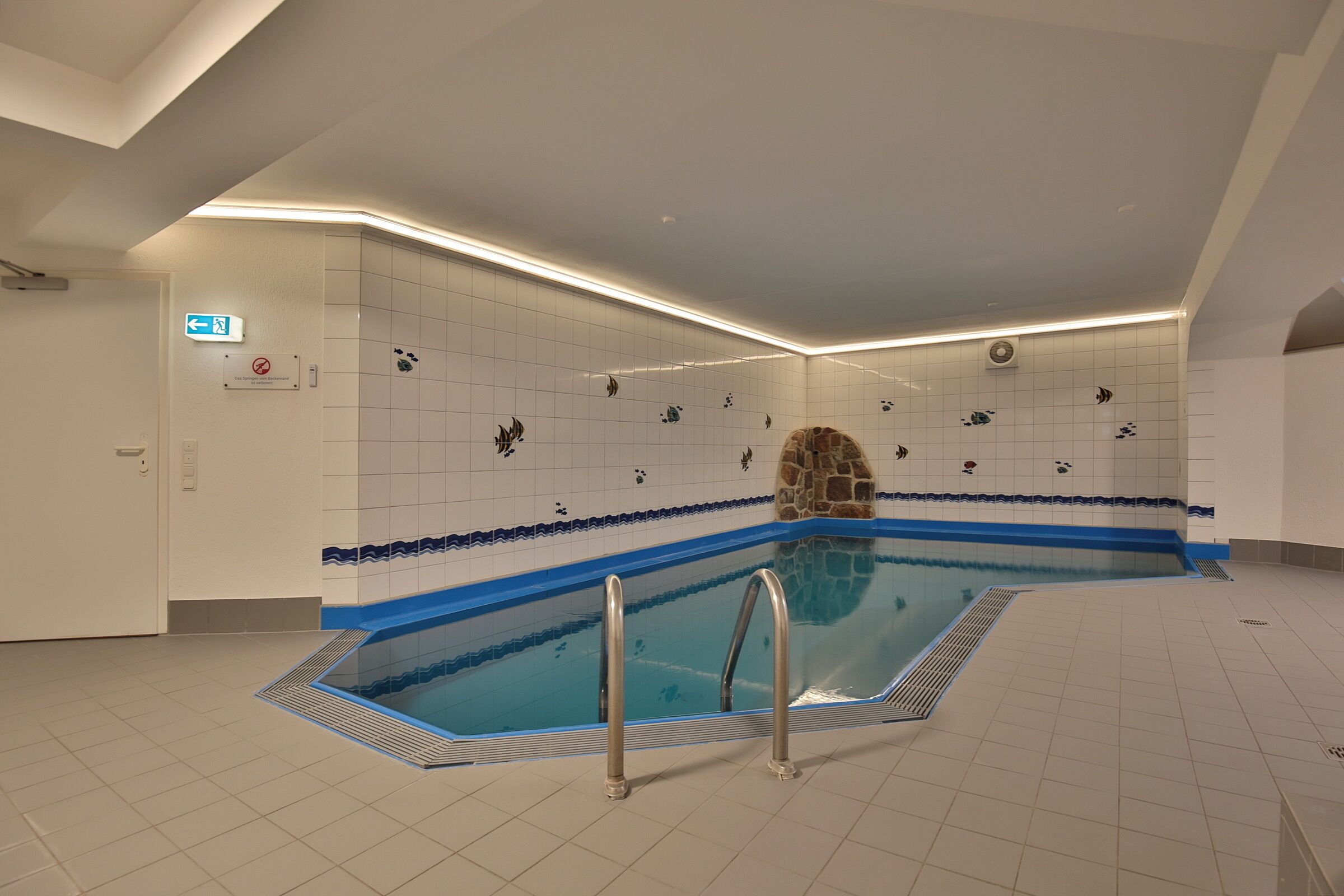 Une piscine intérieure dont un des murs est recouvert de carreaux formant un motif bleu et blanc, avec une arche en pierre et un panneau indiquant qu’il est interdit de fumer dans cet espace.
