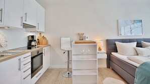 Une cuisine moderne avec des armoires blanches, un four encastré et une machine à café. On y trouve un tabouret de bar blanc et une table de chevet avec une lampe et une petite étagère. Un canapé gris avec des coussins blancs se trouve dans l’espace salon.