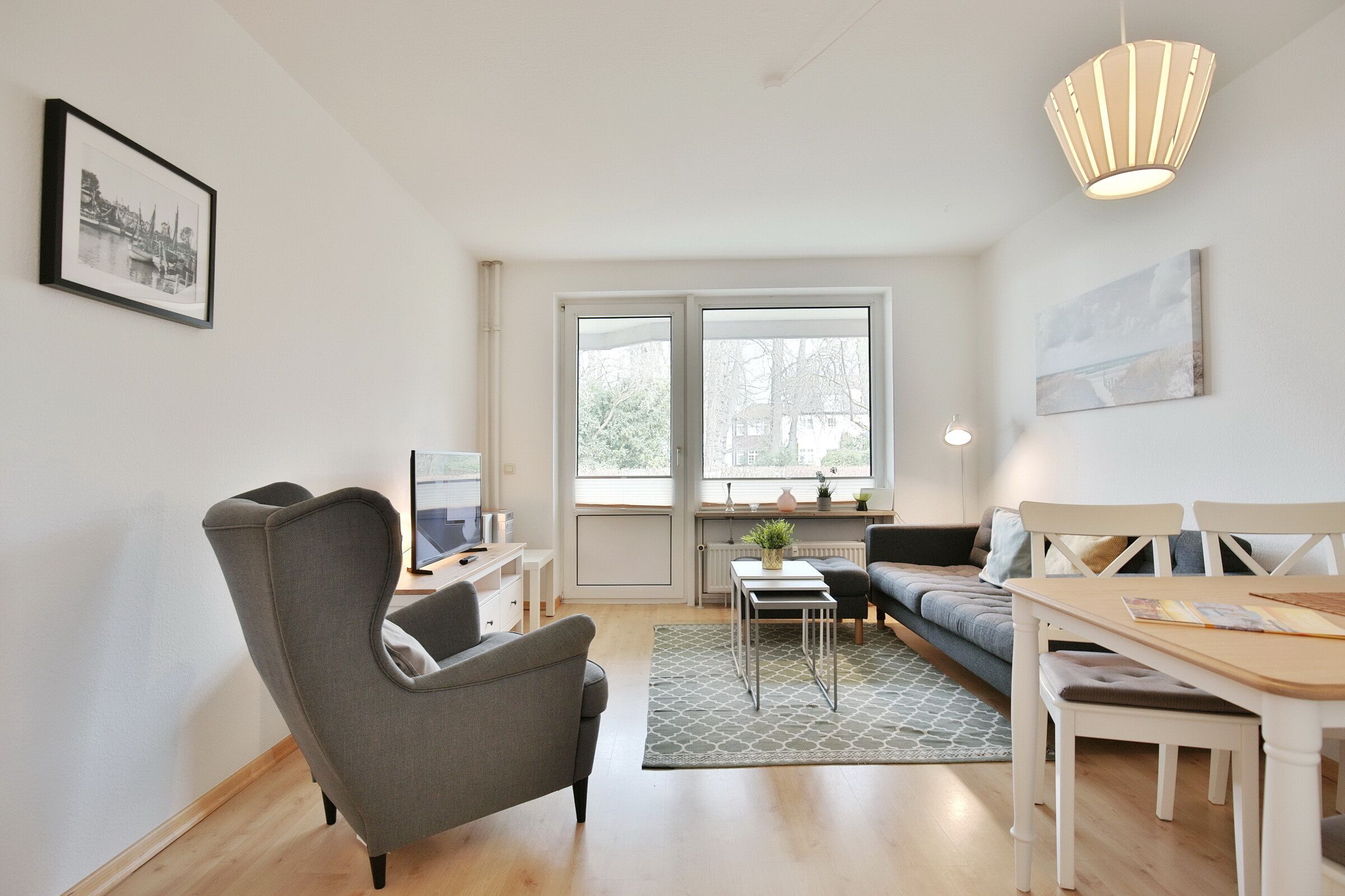 A modern living room with a grey armchair, a sofa, a dining table with chairs, a small side table, and a large window with a view of trees and buildings.