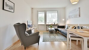 Ett modernt vardagsrum med en grå fåtölj, en soffa, ett matbord med stolar, ett litet sidobord och ett stort fönster med utsikt över träd och byggnader.
