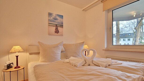 Ett hotellrum med en väl bäddad säng, en tavla på väggen, en lampa på ett sidobord och ett fönster med utsikt över byggnader och träd.