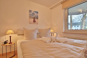 Un habitación de hotel con una cama bien hecha, un cuadro en la pared, una lámpara sobre una mesita de noche y una ventana con vistas a edificios y árboles.