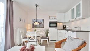 Eine moderne Küche mit weißen Schränken, ein Essbereich mit weißem Tisch und Stühlen sowie ein Korbstuhl mit Kissen.