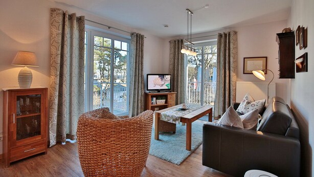 A living room with a sofa, a television, a wooden cabinet, and a wicker chair. - Villa am Meer Strandallee 48 Appartement 10 (Timmendorfer Strand)