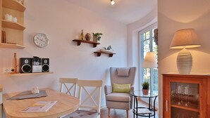 A cozy living room with a round wooden table, two chairs, a sofa, a small side table with a lamp, a cabinet with glass doors, and wall-mounted shelves with decorative items and a clock. - Villa am Meer Strandallee 48 Appartement 10 (Timmendorfer Strand)