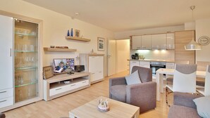 A modern living room with a TV, sofa, and a dining area with chairs. A kitchen is visible in the background.