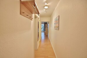 Un couloir étroit avec un plancher en bois et des murs blancs. On y trouve un luminaire au plafond et une porte au bout du couloir.
