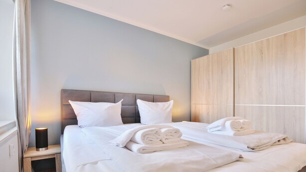A neatly made bed with white linens and towels, a bedside table with a lamp, and a wooden wardrobe.