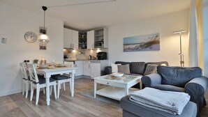 En moderne stue med spisebord, stole, sofa og sofabord. Et køkken ses i baggrunden med skabe og hårde hvidevarer.