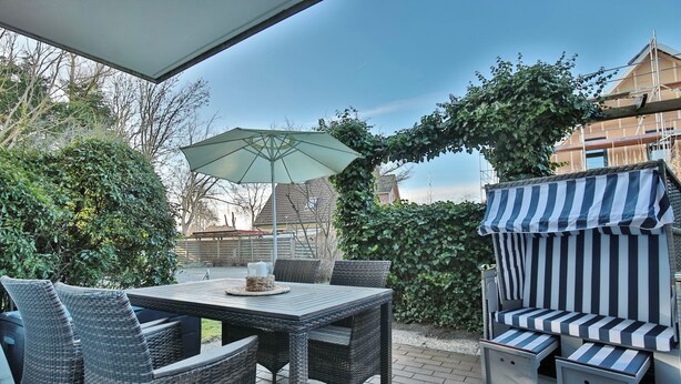 En udendørs terrasse med fletningsmøbler, et bord med en grøn parasol og et stribet markise.