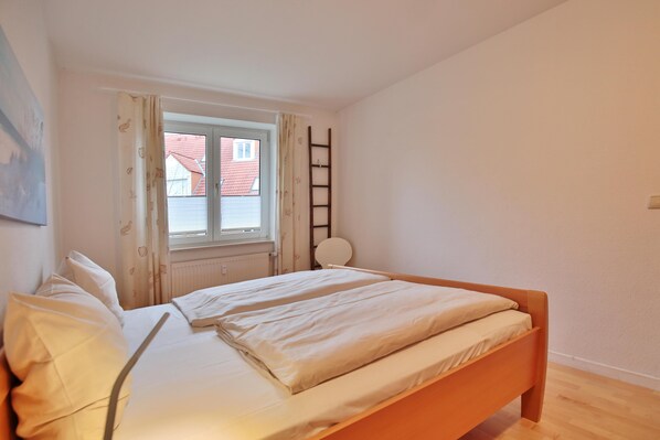 A bedroom with a bed, a window with curtains, a ladder, and a radiator. - Wohnpark am Mühlenteich Appartement 9 (Timmendorfer Strand)