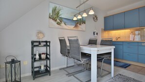A dining area with a white table and chairs, a painting of a beach scene, and blue cabinets in the kitchen area. - Am Elchgrund Appartement 10 (Timmendorfer Strand)