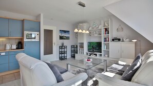 A modern living room with a glass coffee table, white sofas, a television, and a kitchen area with blue cabinets and a microwave. - Am Elchgrund Appartement 10 (Timmendorfer Strand)
