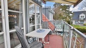 A balcony with a white table and striped chairs, a glass railing, and a view of neighboring houses. - Am Elchgrund Appartement 10 (Timmendorfer Strand)