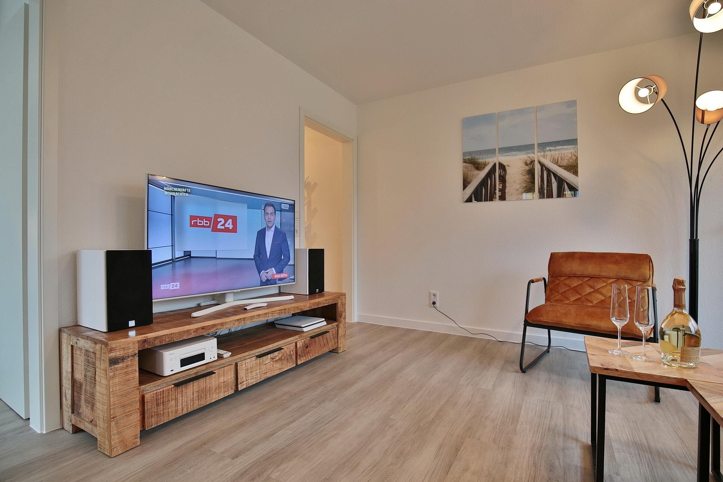 A modern living room with a wooden entertainment unit, a television displaying a news channel, a comfortable armchair, and a table with a bottle and glasses.
