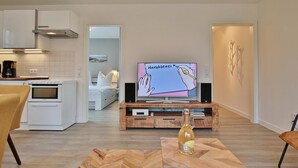 Une cuisine moderne avec des meubles blancs et un four encastré, un coin repas avec une table et des chaises en bois, et un salon avec une télévision et des œuvres d’art décoratives au mur.