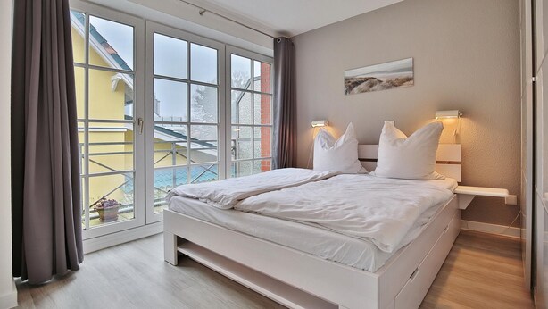 Une chambre avec une grande porte-fenêtre coulissante donnant sur un balcon, un lit avec une literie blanche, une table de chevet avec une lampe et un tableau accroché au mur.
