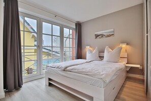 A bedroom with a large sliding glass door leading to a balcony, a bed with white bedding, a nightstand with a lamp, and a painting on the wall. - Haus Parkblick Appartement 6 (Timmendorfer Strand)