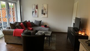 A modern living room with a sectional sofa, a glass coffee table, and a TV on a stand. There are two paintings on the wall and a window with a view of a wooden deck. - Haus Parkblick Appartement 3 (Timmendorfer Strand)
