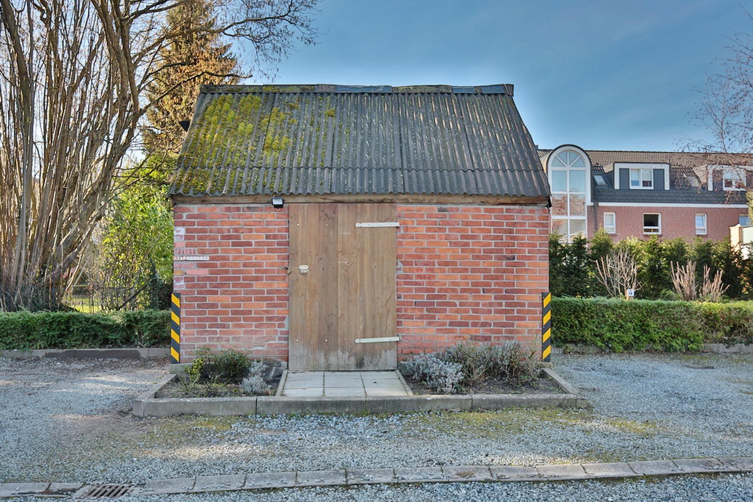 A small brick building with a wooden door, a metal roof, and a gravel driveway.