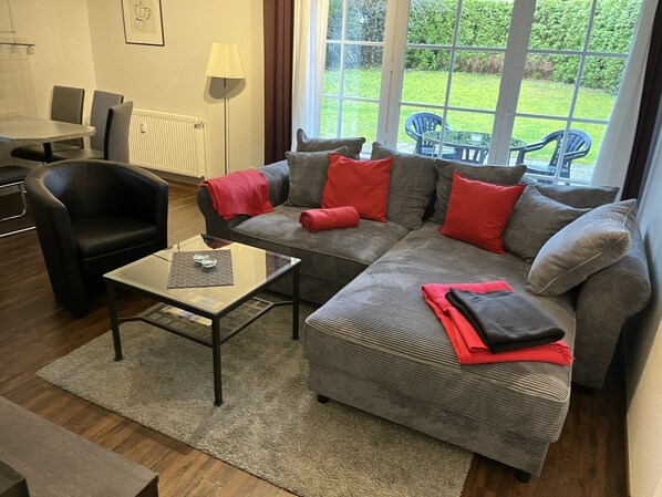 Ein modernes Wohnzimmer mit grauem Sofa, schwarzem Sessel und einem Glastisch. Große Fenster bieten Blick auf einen Garten.
