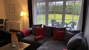 A living room with a sofa set, a coffee table, and a lamp. There is a view of a garden through large windows. - Haus Parkblick Appartement 3 (Timmendorfer Strand)