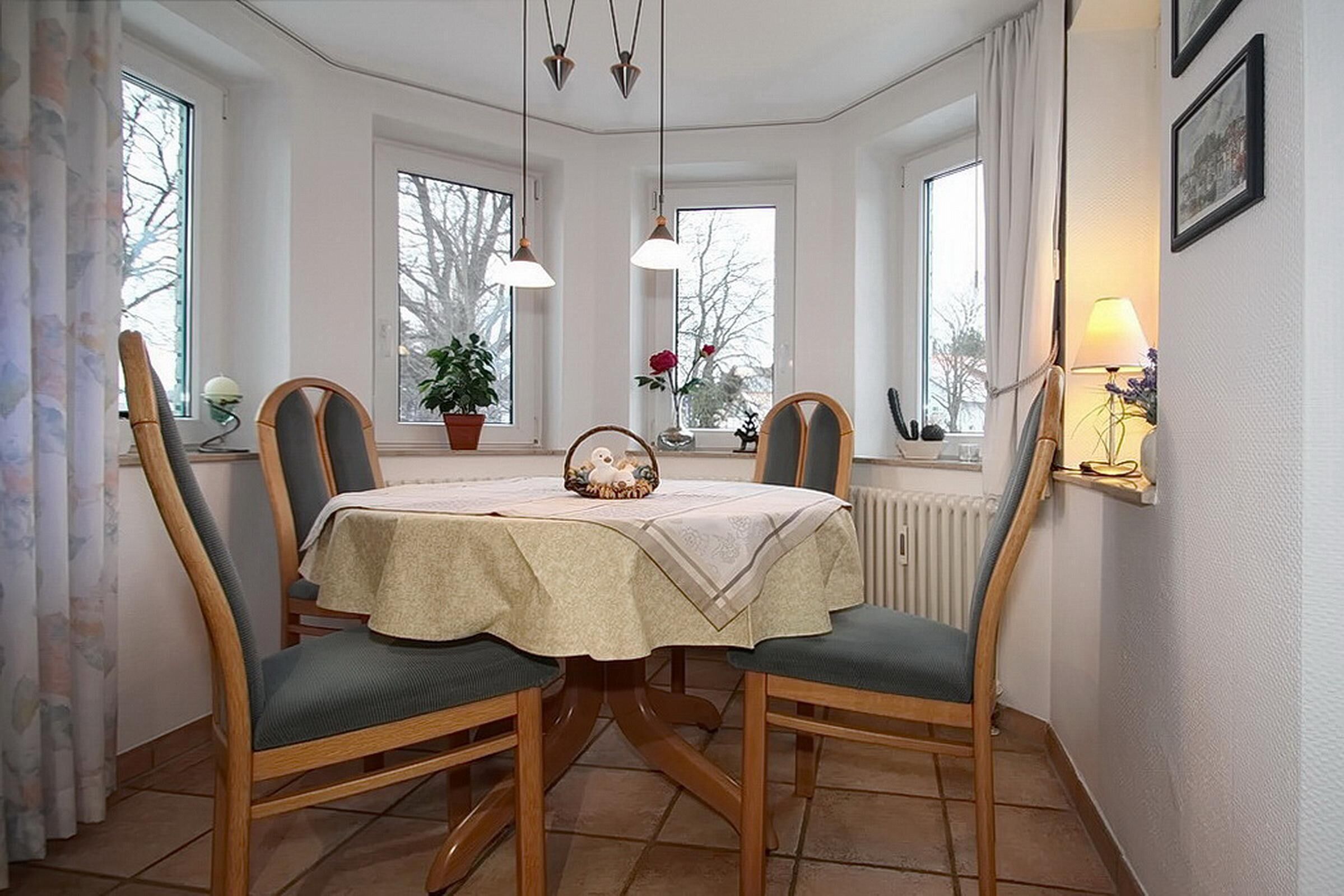 A dining room with a table set for four, surrounded by wooden chairs with green cushions. There are three pendant lights hanging above the table, and the room has large windows with curtains.