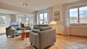 Un salon spacieux avec un canapé gris, une table basse en bois et un téléviseur sur le côté gauche. De grandes fenêtres, ornées de rideaux, laissent entrer la lumière naturelle et offrent une vue sur l’extérieur.