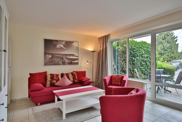 Un salon moderne avec un canapé rouge, une table basse blanche et une grande fenêtre donnant sur un paysage verdoyant et un immeuble.