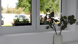 A white console table with a white vase containing green plants. Two windows with black frames are visible, showing a parking lot and trees outside.