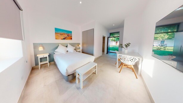 A modern bedroom with a bed, bedside tables, a desk, and a chair. There is a painting on the wall and a window with a view of greenery. - One Bedroom Apartment (Toledo)