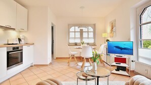 Een moderne keuken met witte kasten en een inbouwoven, een eethoek met een ronde tafel en stoelen, een flatscreen televisie op een standaard, en een groot raam met uitzicht op groen.