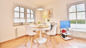 Een moderne eetruimte met een ronde tafel, witte stoelen, een tv op een dressoir en een raam met zonwering.