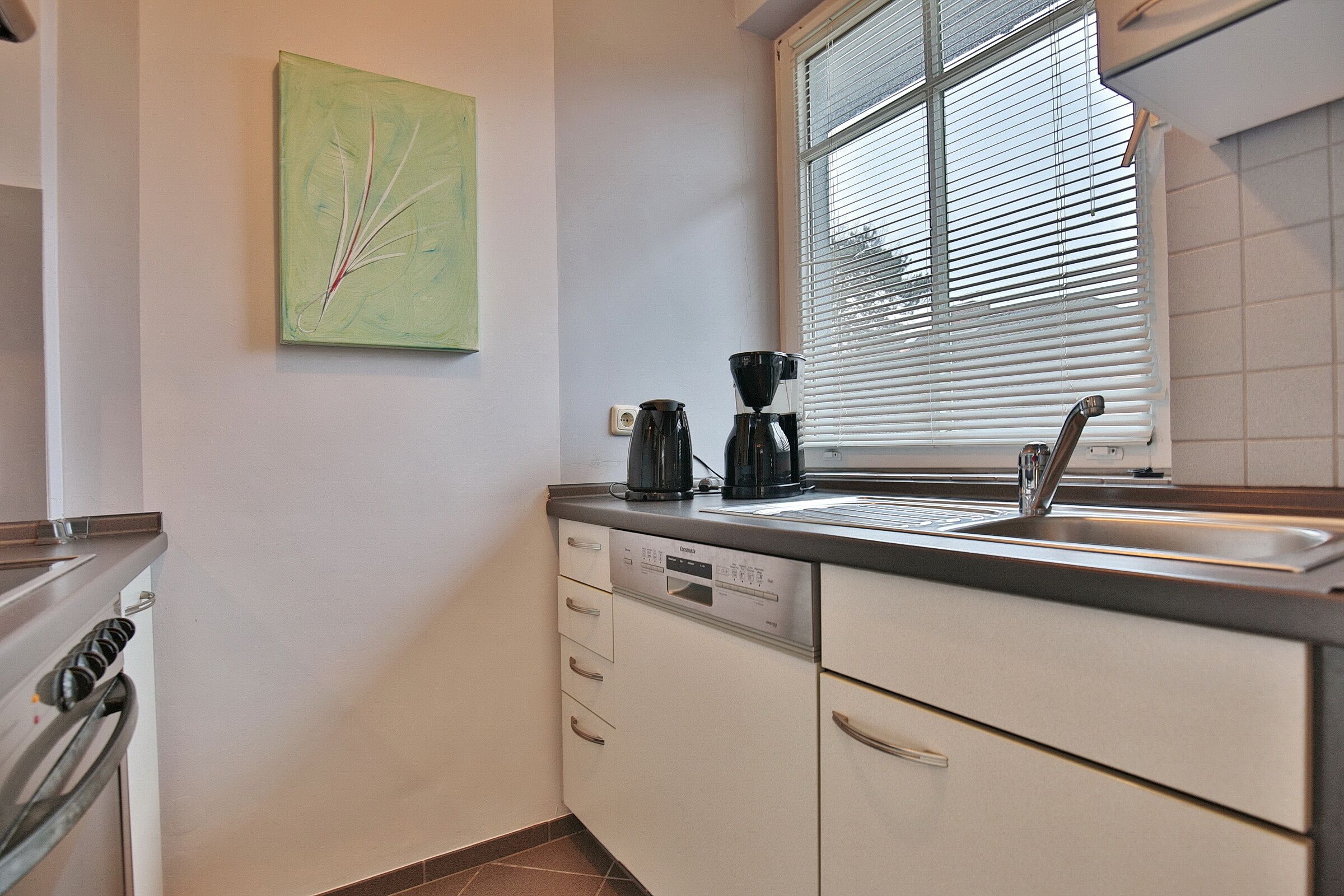 Een moderne keuken met een vaatwasser, koffiezetapparaat en een raam met jaloezieën.