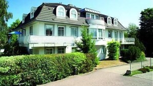 Een groot, meerlaags huis met een opvallend balkon op de tweede verdieping, omgeven door groen.