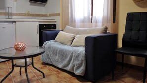 A living room with a sofa, a small round table, and a chair. There is a television on the wall and a window with curtains.