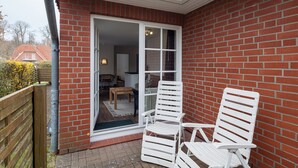 Un patio con dos sillas blancas y una mesa de madera, un muro de ladrillo y un ventanal corredero que da a un espacio interior con una lámpara y una silla.