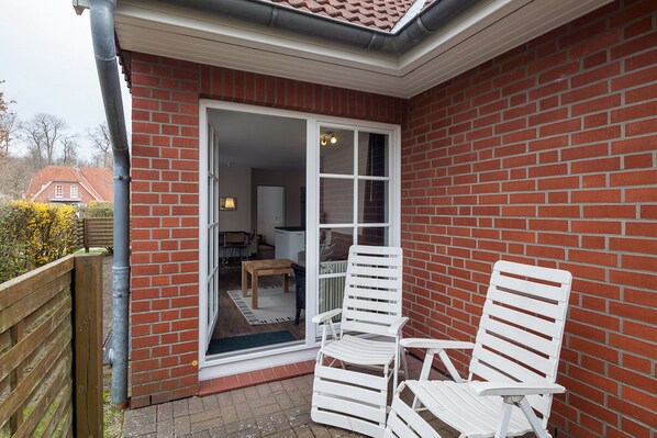 A patio with two white chairs and a wooden table, a brick wall, and a sliding glass door leading to an interior space with a lamp and a chair.