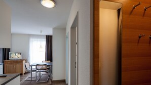 Habitación de hotel con puerta de madera, un pequeño comedor con mesa y sillas, un televisor sobre un mueble de madera y una lámpara en el techo.