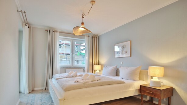 Un dormitorio con cama, mesitas de noche, lámpara, ventana con cortinas y vista a un edificio.