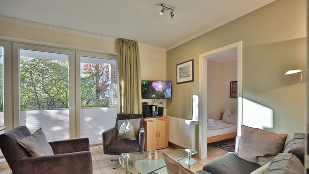 A modern living room with a sofa, armchair, and glass coffee table. A television and speakers are on a wooden cabinet. The room has large windows with a view of trees. - Moderne Strandvilla Appartement 2 (Timmendorfer Strand)
