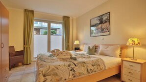 A bedroom with a bed, a nightstand, a chair, a window with curtains, and a painting on the wall. - Moderne Strandvilla Appartement 2 (Timmendorfer Strand)
